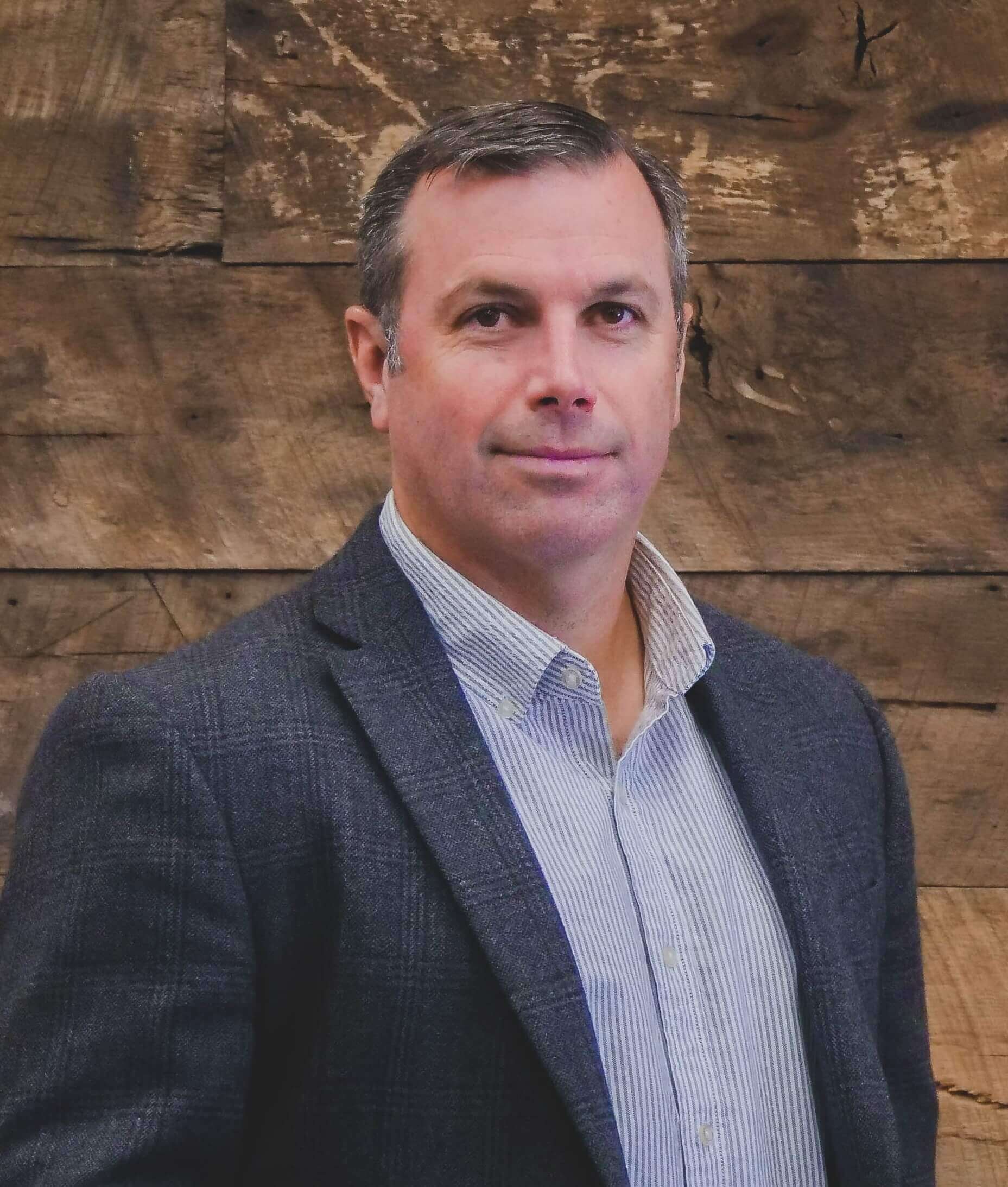 Mr. Madison Burt stands before a wooden accent wall wearing a sport coat and collared shirt