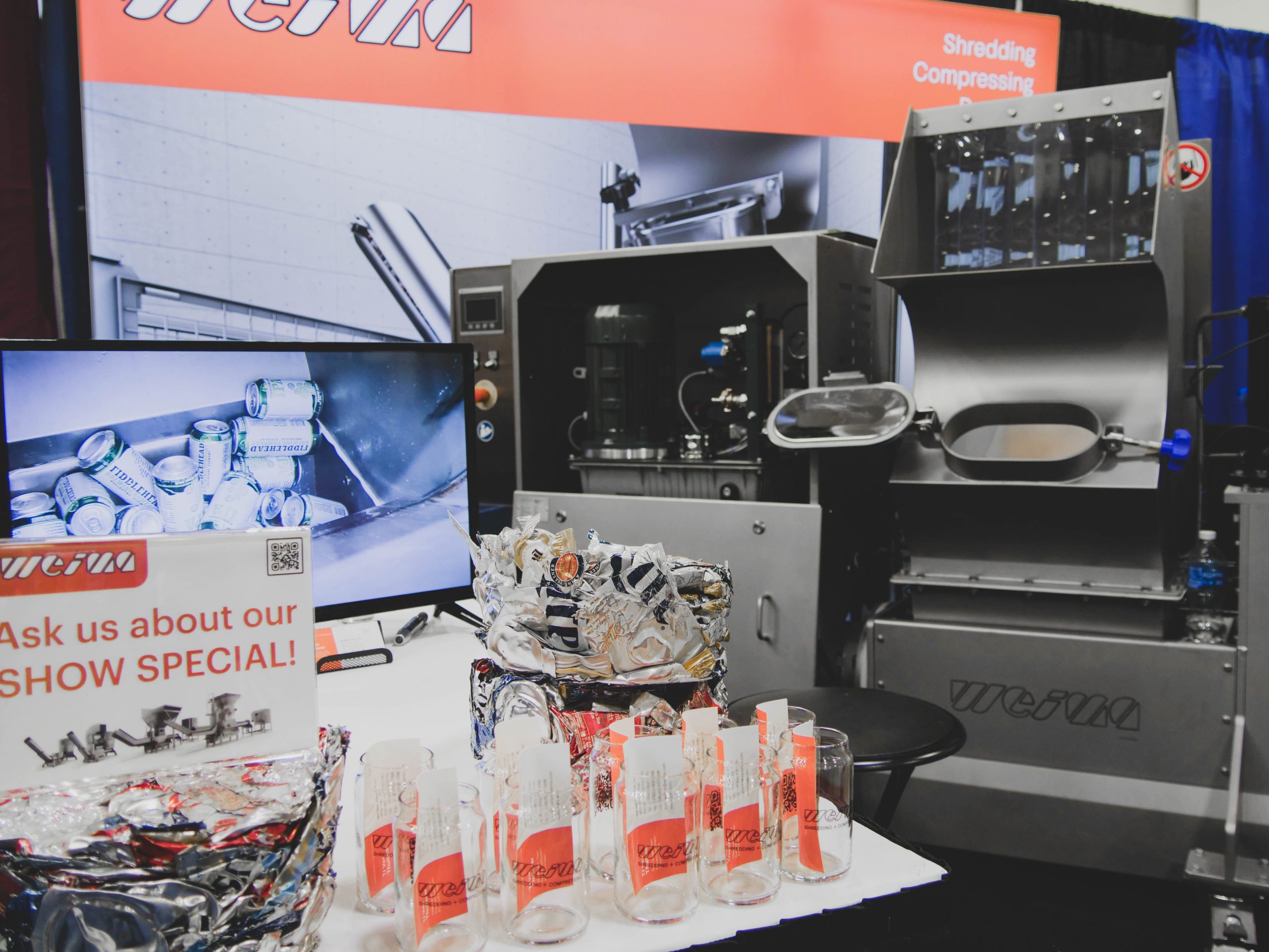 A WEIMA E.200 machine is in a tradeshow booth with a TV and some glasses for giveaways.