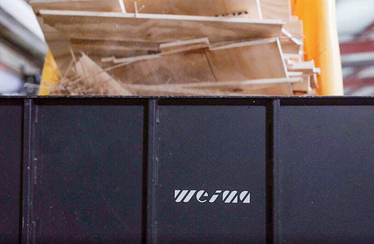 A load of wood scrap is being dumped into a gray WEIMA shredder hopper.