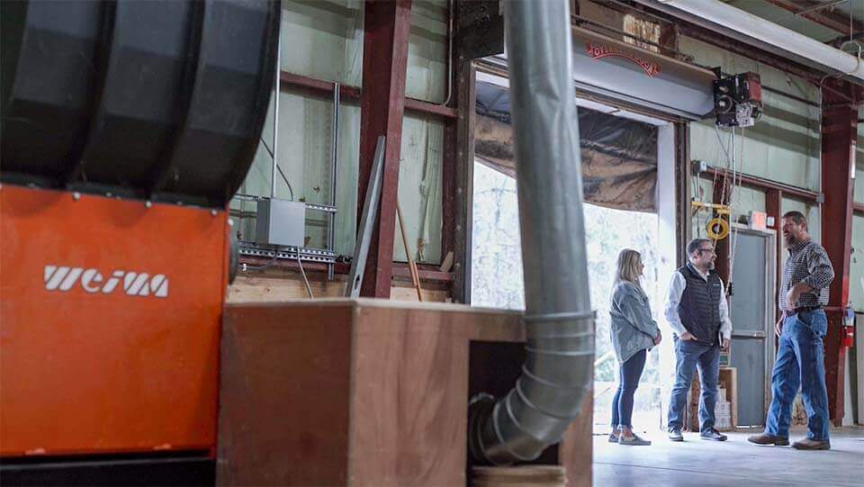 Three people are standing and speaking with each other in a warehouse next to an orange WEIMA shredder.