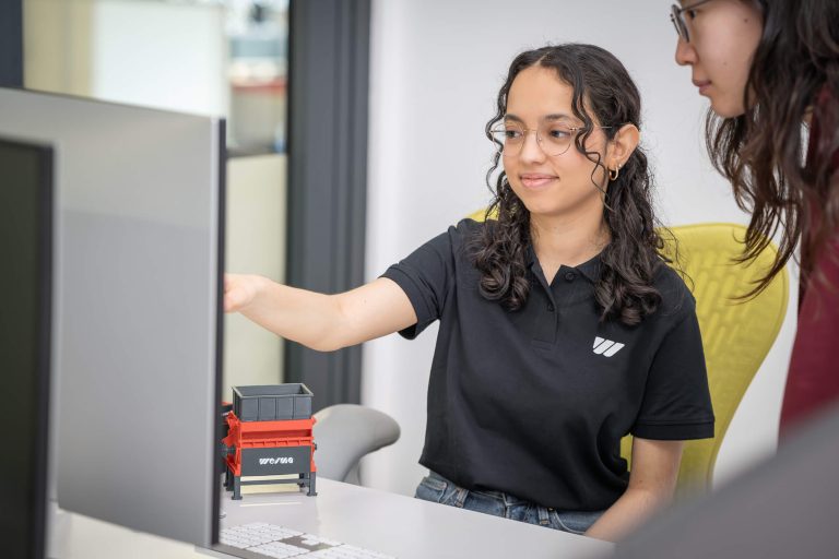Mitarbeiterin mit Brille und schwarzem Shirt sitzt vor einem PC und zeigt auf den Bildschirm, daneben ein 3D-Druck Modell eines WEIMA Shredders