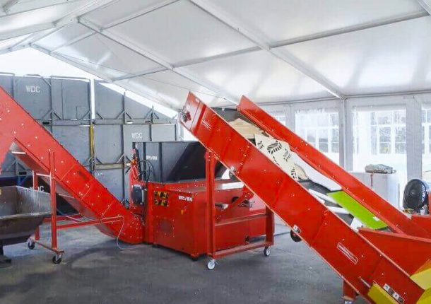 A red shredder machine is connected to an infeed and discharge conveyor. Pieces of wood waste are on the infeed conveyor. A black bin waits at the end of the discharge conveyor to collect the shredded material.