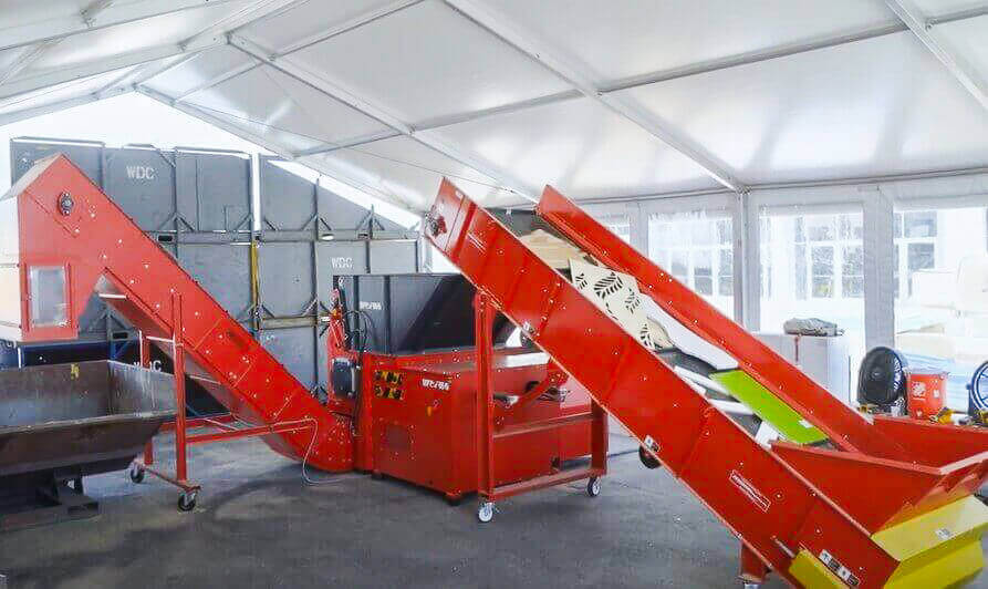 A red shredder machine is connected to an infeed and discharge conveyor. Pieces of wood waste are on the infeed conveyor. A black bin waits at the end of the discharge conveyor to collect the shredded material.