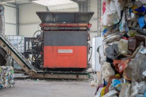 Ein orangefarbener Shredder in einer Industriehalle mit Post-Consumer Müll im Vordergrund
