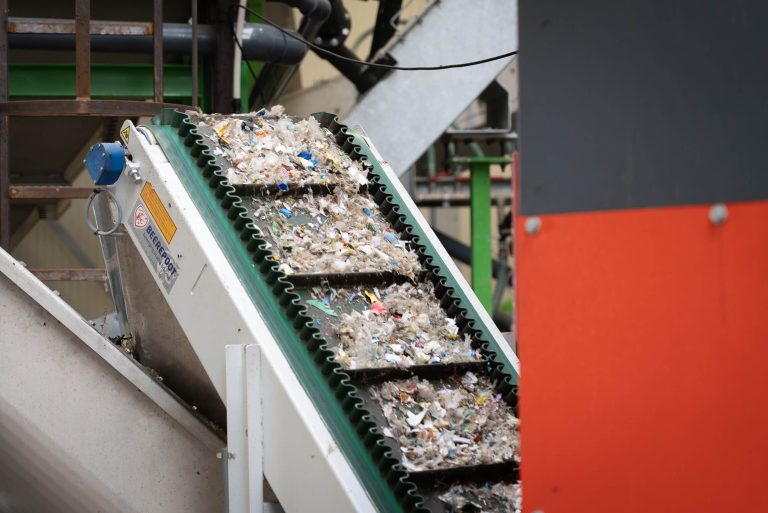 A green conveyor belt with shredded colorful post-consumer plastic with an orange shredder in the foreground