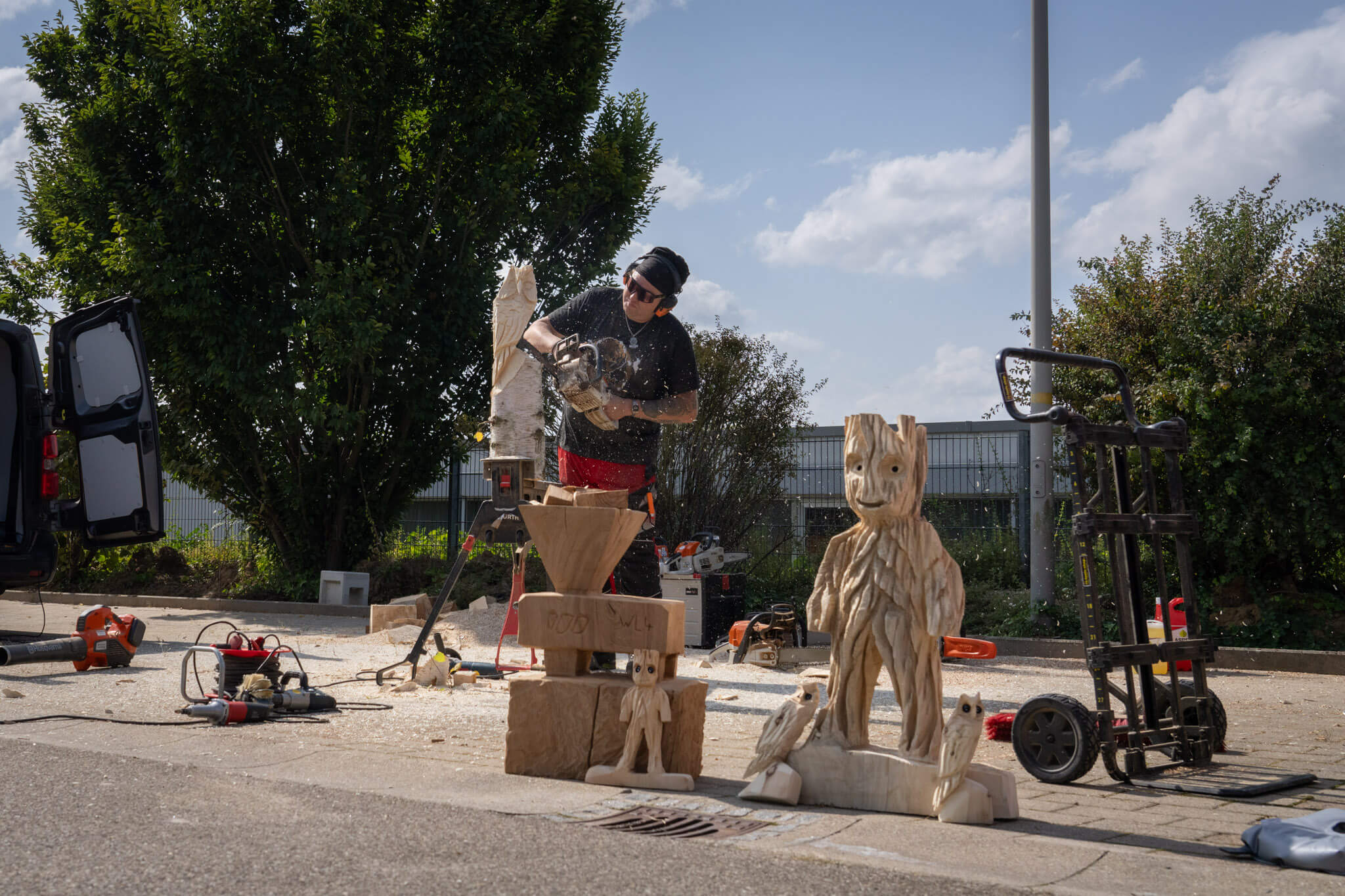 A man carves figures out of a block of wood with a chainsaw. In the foreground are already finished figures
