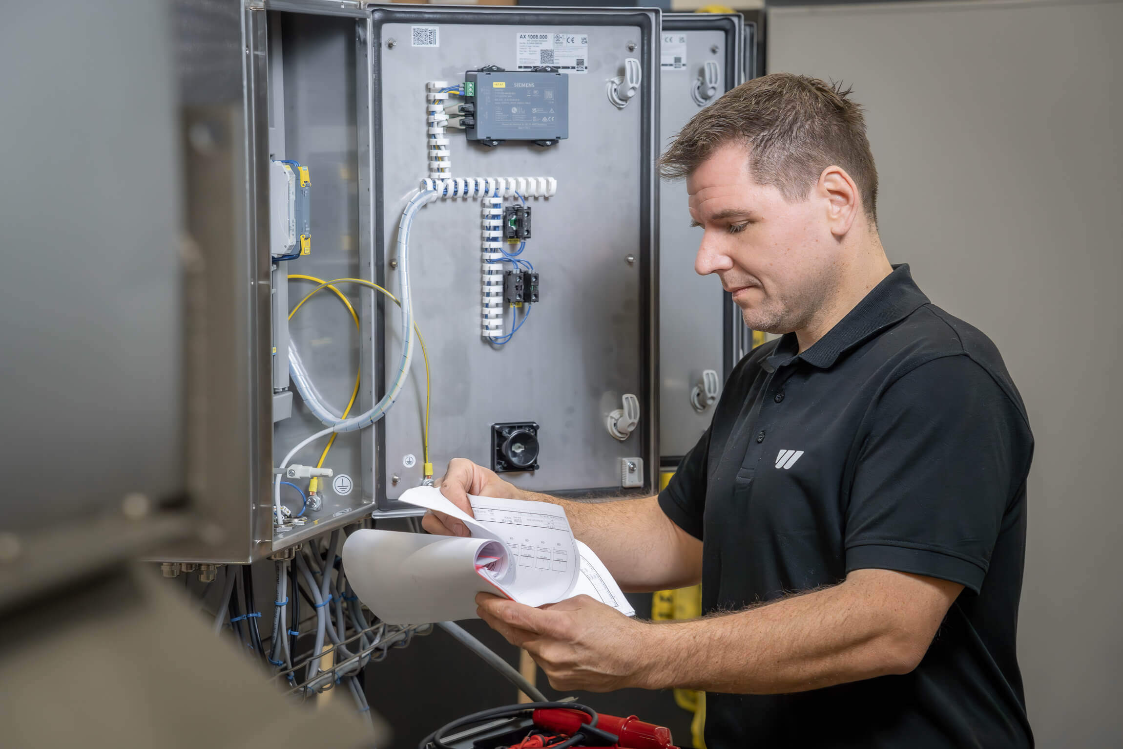 Man steht vor einem offenen Schaltschrank und schaut auf einen Plan vor sich