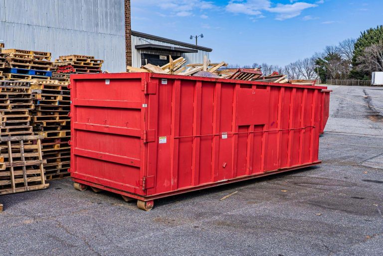A red dumpster behind a warehouse is filled with wood scraps. Next to it is a stack of wooden pallets.