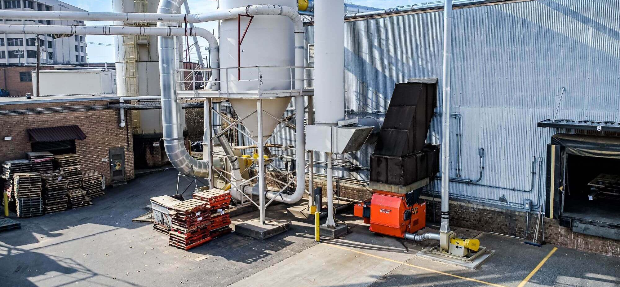 An orange WEIMA shredder is positioned behind a warehouse, surrounded by an air system with a white silo and metal duct work.