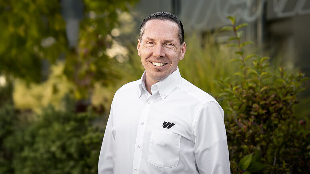 Patrick Henzler, Sales Director at WEIMA, in front of a green plant background with a white shirt, smiling