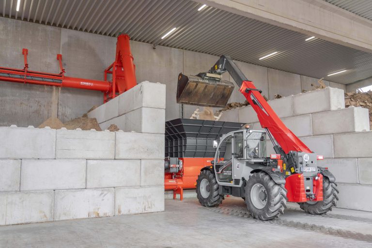 A telescopic loader transports wood scraps into a large hopper for further processing.