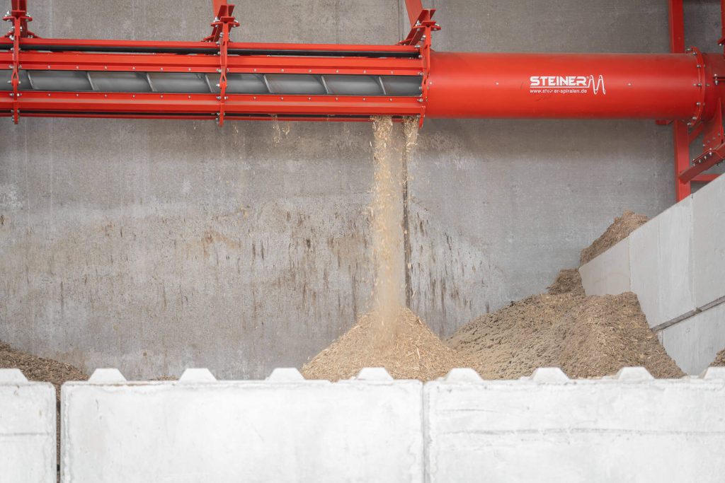 Shredded wood chips fall from a red Steiner spiral conveyor into a concrete bunker in the Trier firewood plant.
