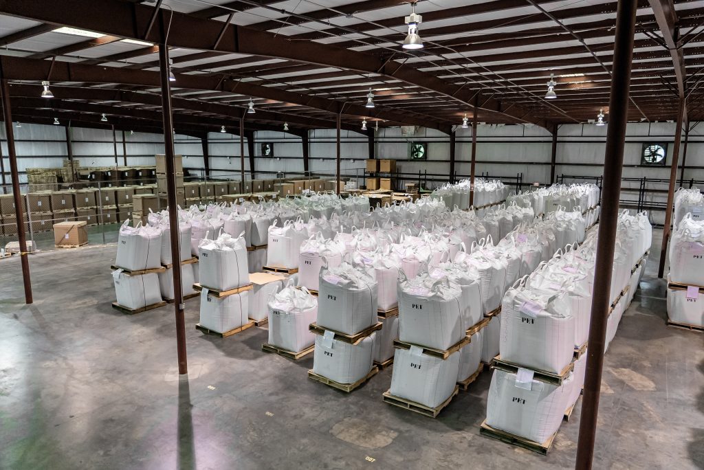Numerous big bags labeled “PET” are neatly stacked on pallets in a large industrial hall.