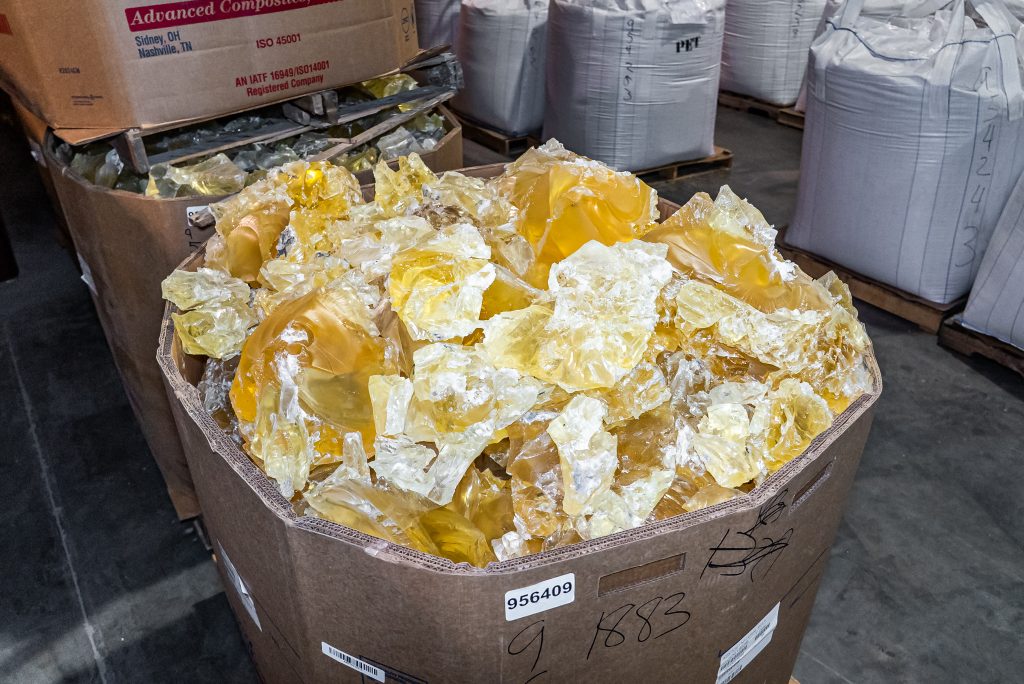 A large cardboard container is filled with yellow, transparent chunks of plastic and stands in the middle of other big bags in a warehouse.