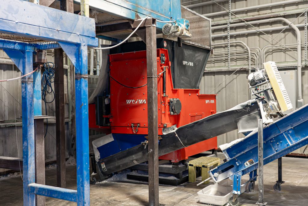 An industrial WEIMA shredding plant with feed hopper and blue conveyor belts is located in a production hall for plastics processing.