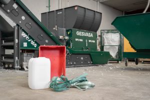 Ein weißer Kanister, eine rote Kunststoffbox und grüne Kunststoffbänder vor einem grünen Shredder für das Recycling