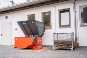 Ein orangefarbener Shredder vor einem weißen Gebäude mit einer Kiste mit alten Fassdauben