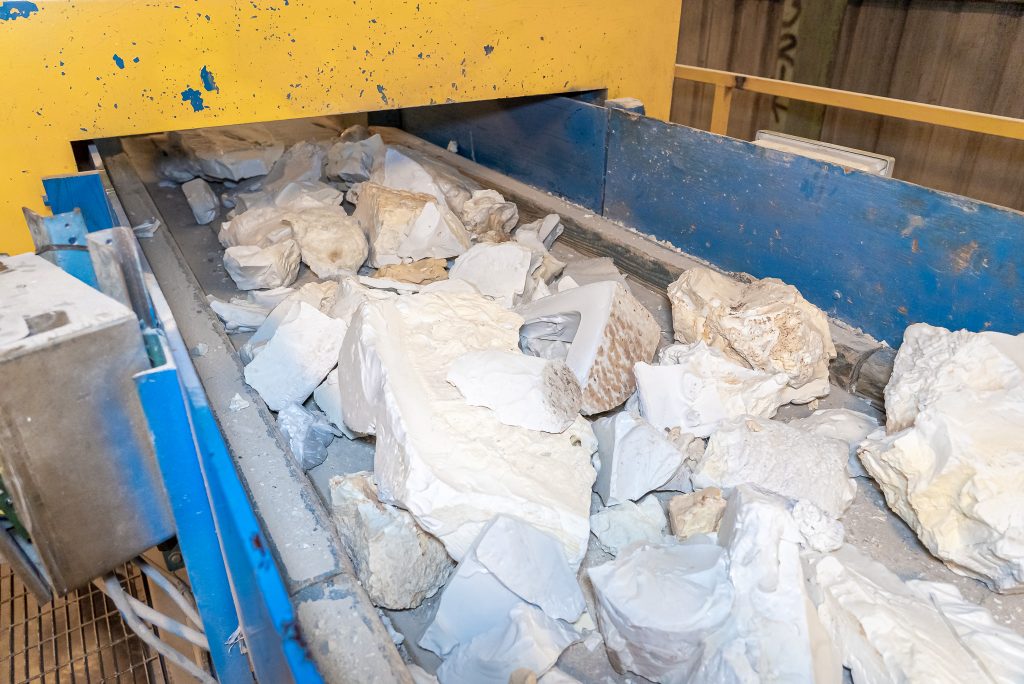 Large chunks of white and beige material lie on a conveyor belt and are transported to a yellow industrial plant for further processing.