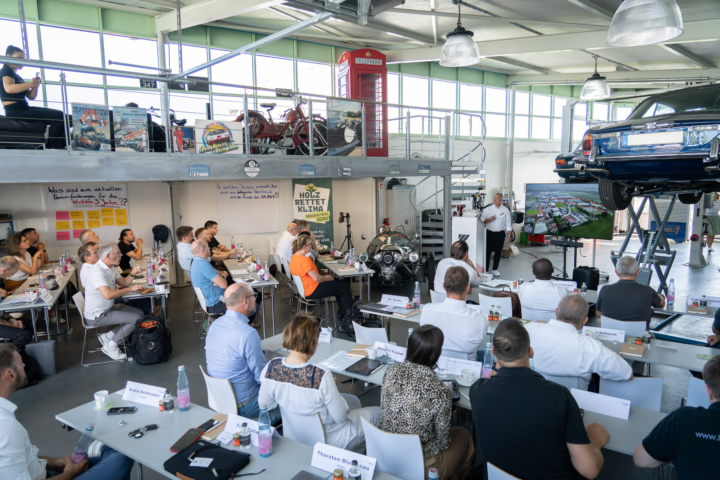 A man gives a lecture on waste wood management, standing in the middle of old vintage cars from the WEIMA garage