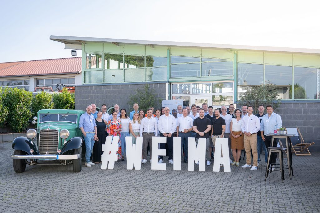 A group of people are standing in front of a building. In the foreground are white letters with the lettering #WEIMA, next to them is a green vintage car.