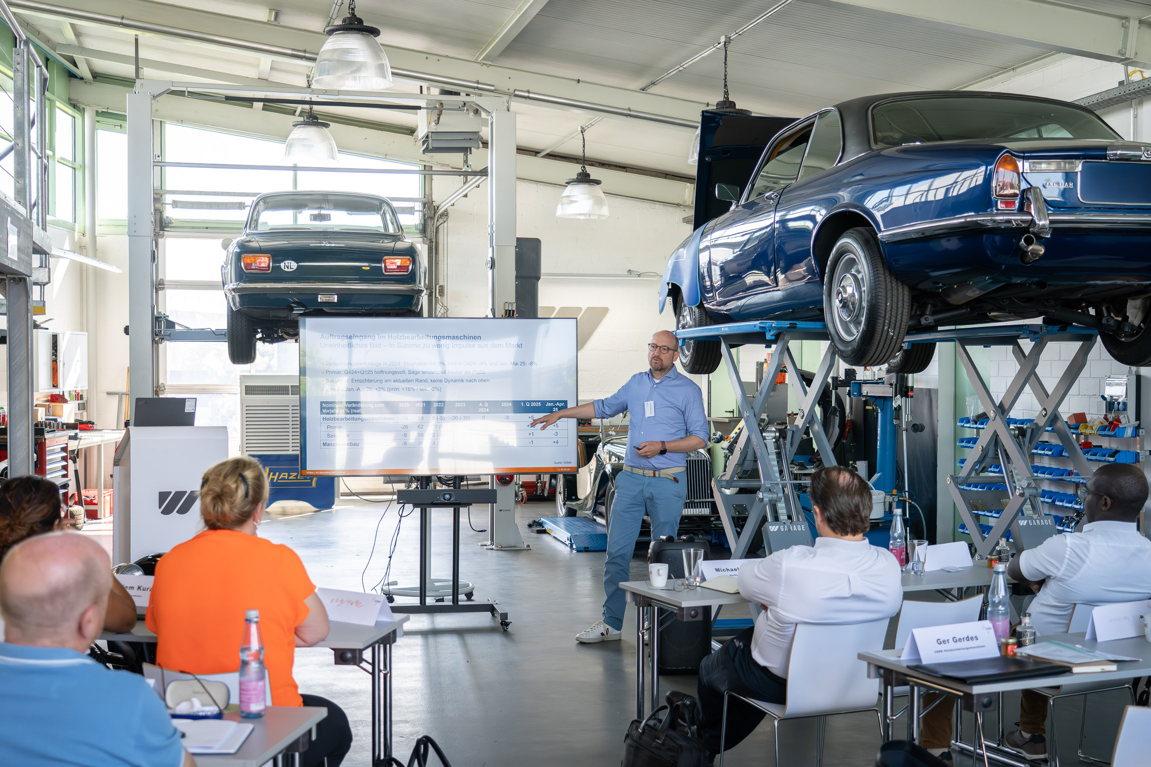 A man gives a lecture on the current situation in the timber market, standing in the middle of old vintage cars from the WEIMA garage