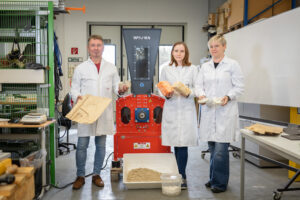 Three researchers in white coats standing in front of an orange shredder with various exhibits in their hands.