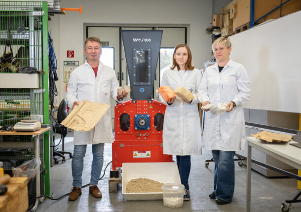 Three researchers in white coats standing in front of an orange shredder with various exhibits in their hands.